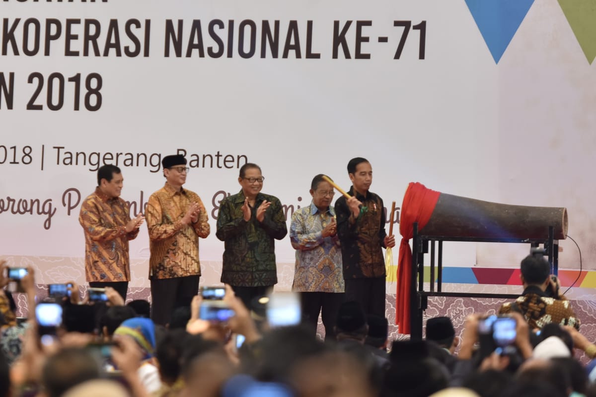 Presiden Joko Widodo memukul kentongan di atas panggung Hari Koperasi Nasional ke-71, didampingi empat pejabat berbatik, disaksikan hadirin yang merekam dengan telepon genggam.